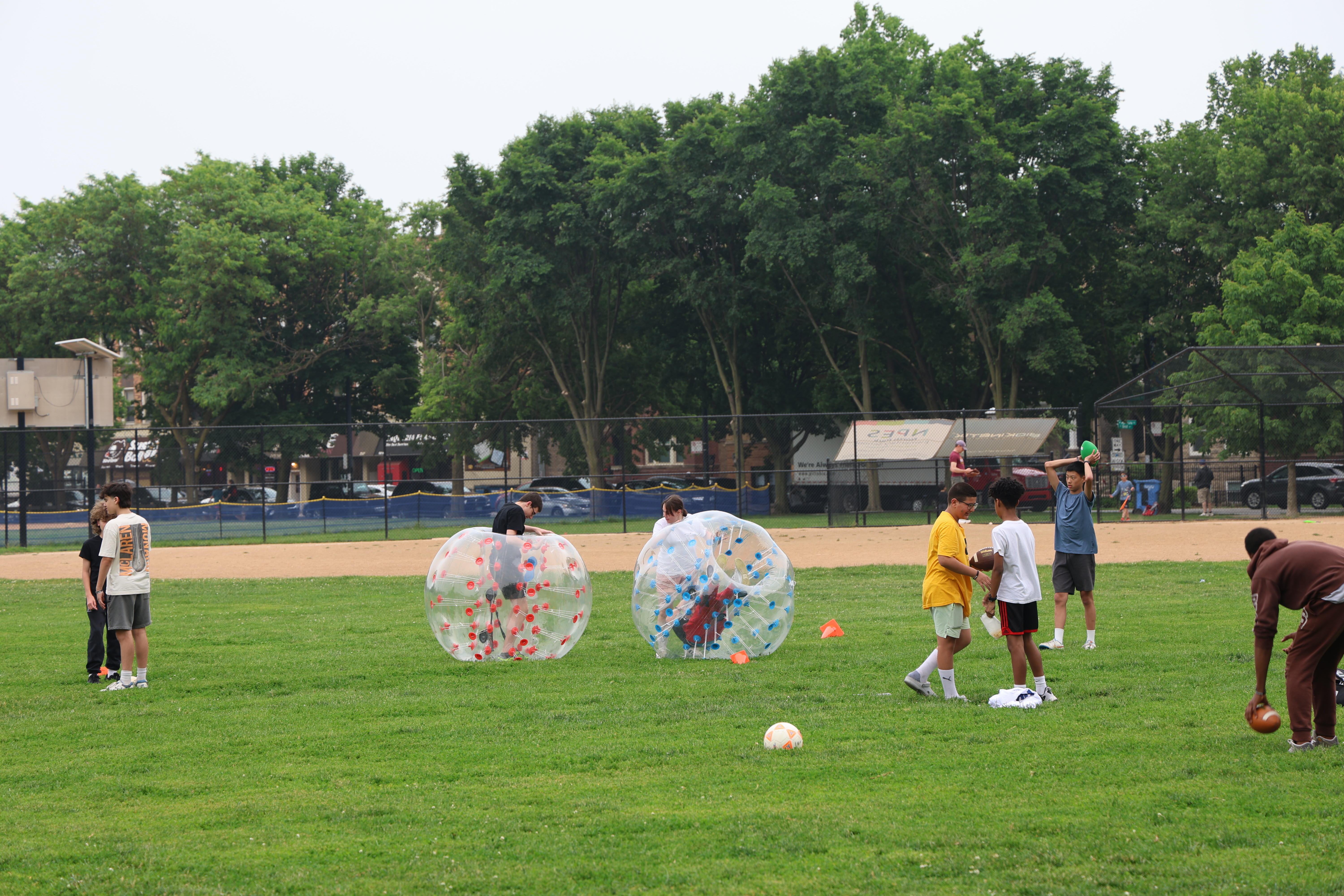 Kids playing various sports on a grassy field. Some kids inside inflatable bumper balls.